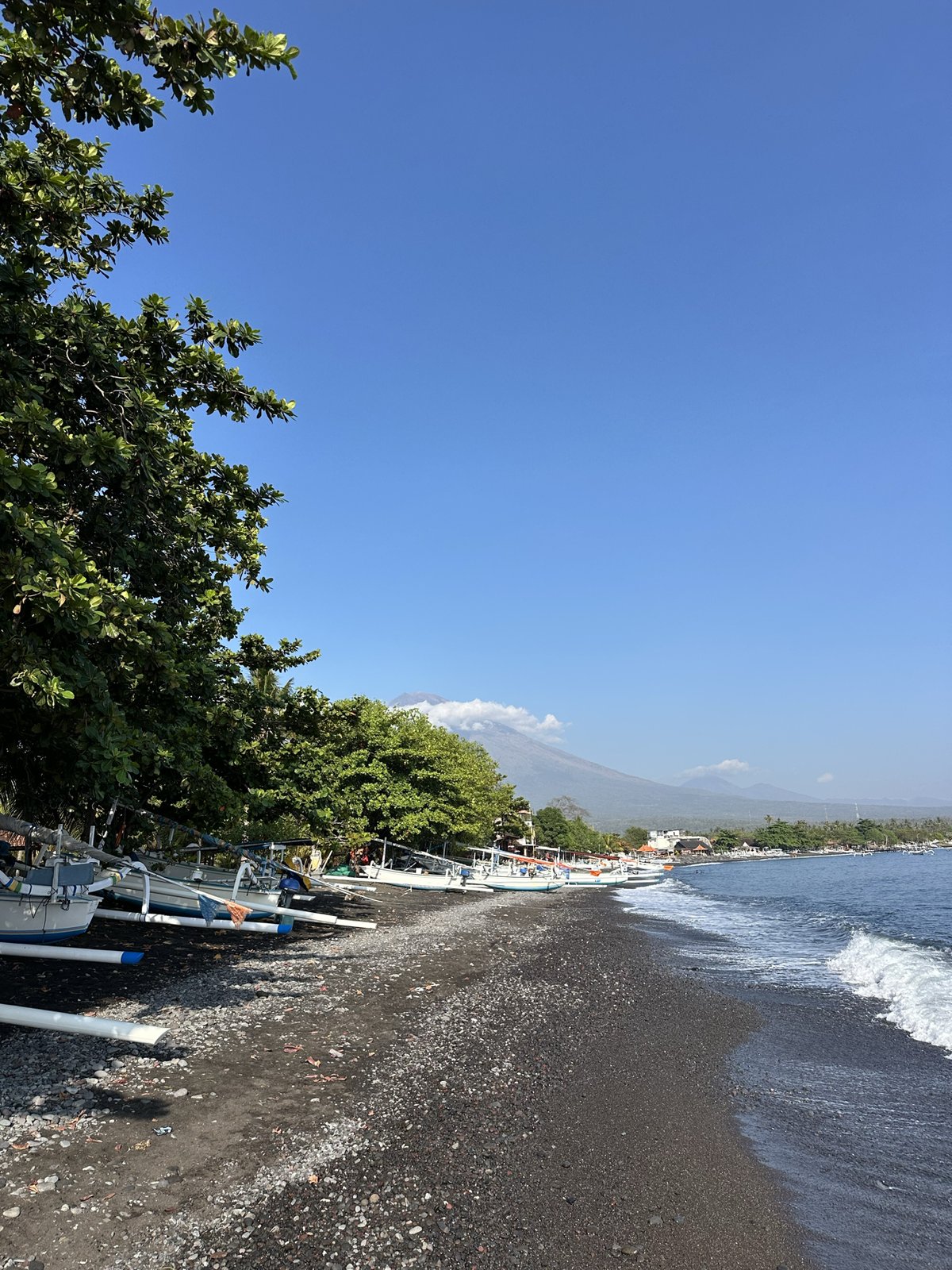 Vague sur les galets noirs d'Amed, jukungs et arbres sous le Mont Agung