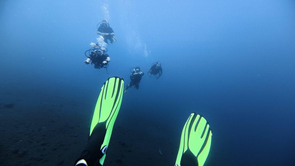 Plongeurs en formation sous l'eau, palmes jaune fluo au premier plan, fond de sable noir à Amed